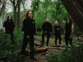 Cinq hommes en noir posent dans une forêt. Le groupe, qui pourrait être Cattle Decapitation, se tient parmi les arbres et la végétation luxuriante.