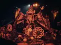 Un personnage masqué, drapé dans des tissus ornés, semble présider un rituel. Autour de lui, des offrandes de fruits, des serpents stylisés et des symboles sacrés évoquent un culte du feu mystérieux.