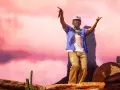 Tyler Okonma, micro en main, danse sur scène devant un décor rose pastel. Il porte une casquette, une chemise bleue ouverte sur un t-shirt blanc et un pantalon beige.