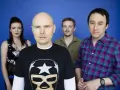 Les membres de The Smashing Pumpkins posent devant un fond bleu. Billy Corgan porte un t-shirt noir avec un masque de lucha.