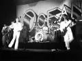 Le groupe Trust en concert, avec le chanteur en blanc au centre, un guitariste à gauche et un autre guitariste à droite. La scène est décorée d'une grande bannière avec le logo du groupe.