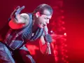 Till Lindemann, chanteur du groupe metal allemand Rammstein, en pleine performance sur scène, micro à la main, sous un éclairage rouge intense.