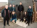 Les membres de Dropkick Murphys posent devant un mur sombre, certains accroupis, d'autres debout, prêts pour leur prochain concert.