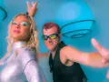 Un groupe hollandais des années 90, avec une femme au maquillage bleu vif et un homme aux lunettes futuristes, pose devant des sphères lumineuses bleues.
