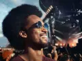 Homme souriant à un concert, portant des lunettes de soleil et un bouchon d'oreille pour protéger son audition.