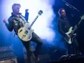 Deux membres de Queens of the Stone Age en pleine performance, l'un jouant de la guitare, l'autre de la basse, sous des éclairages bleus intenses.