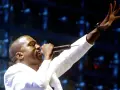 Kanye West, aux origines de sa carrière, chante sur scène, micro à la main, bras levé.