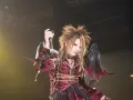 Un membre de X Japan, cheveux ébouriffés, maquillage prononcé, costume sombre et ailes de démon, chante sur scène.
