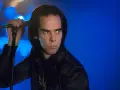 Nick Cave, chanteur du groupe australien des années 80, sur scène, micro à la main, regard intense sur fond bleu.