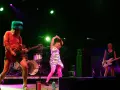 Sur scène, le groupe Bikini Kill enflamme le public. Une guitariste porte un chapeau vert fluo, une autre joue de la basse, et une troisième danse avec énergie.