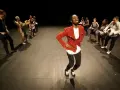 Un danseur afro 93, en veste rouge, mène une troupe sur scène. Les danseurs, aux tenues variées, exécutent des mouvements synchronisés.