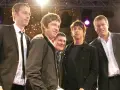 Cinq hommes, dont le groupe Oasis, posent ensemble sur scène, souriants sous les lumières.