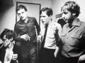 Les membres de Joy Division, Ian Curtis, Bernard Sumner, Peter Hook et Stephen Morris, posent ensemble, certains fumant, d'autres tenant des boissons.