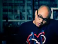 Un homme chauve avec des lunettes, parlant au téléphone, porte un t-shirt avec un logo coloré. Il pourrait bien être celui qui a inventé le rap.