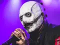 Le chanteur Slipknot, masqué et terrifiant, tient un micro sur scène sous une lumière violette.
