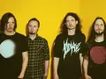 Quatre hommes aux cheveux longs posent devant un fond jaune vif. Leurs t-shirts portent des graphismes distinctifs, évoquant l'énergie brute du magma et la puissance de Gojira.