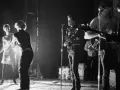 Scène de concert des membres de The Velvet Underground. Une femme danse à gauche, tandis que Lou Reed chante au micro.
