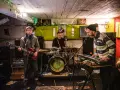 Trois musiciens jouent dans un local sombre, éclairé par des guirlandes. Un guitariste, un batteur et un claviériste, oi boys !