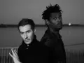 Les membres de Massive Attack, 3D et Daddy G, posent dos à dos, regardant dans des directions opposées, dans une ambiance sombre et contemplative.