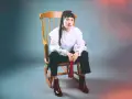 Scout Niblett, assise sur un fauteuil à bascule, porte une blouse blanche à volants et un pantalon noir. Elle tient un pompon rouge.