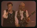 Une chanteuse et un guitariste, peut-être les ramoneurs de menhirs, sur scène. L'homme porte une couronne et une tenue excentrique.