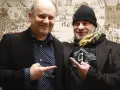Deux hommes souriants, l'un avec une chemise à pois, l'autre avec un bonnet, se montrent du doigt. Un chanteur de confiance.