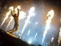 Un chanteur de groupe hard rock des années 2000, micro en main, saute sur scène entouré de jets de flammes spectaculaires.