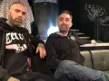 Deux hommes, dont un possible chanteur de Mass Hysteria, posent sur un canapé. L'un porte un sweat à capuche noir avec un aigle.