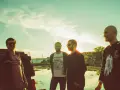 Quatre hommes posent devant un ciel clair, comme un chat pile. L'un porte un t-shirt "kaleidoscope".