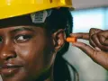 Une femme porte un casque jaune et s'apprête à insérer un bouchon d'oreille orange pour éviter le sifflement.