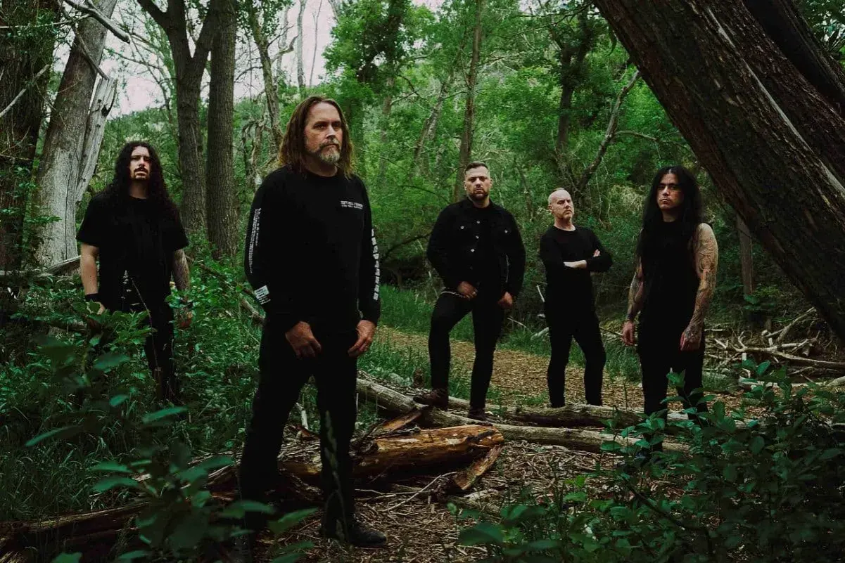 Cinq hommes en noir posent dans une forêt. Le groupe, qui pourrait être Cattle Decapitation, se tient parmi les arbres et la végétation luxuriante.