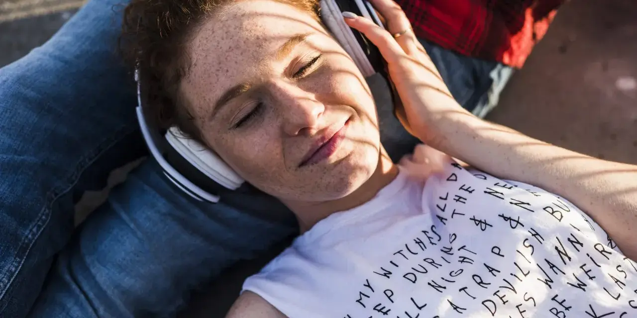 Jeune femme aux taches de rousseur, écouteurs blancs, profitant de la musique. Le volume est à 95 db équivalent, elle semble détendue.