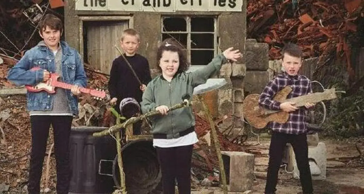 Quatre enfants jouent de la musique devant un panneau "the cranberries". L'un tient une guitare rose, un autre une guitare en bois.
