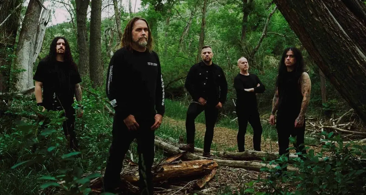 Cinq hommes en noir posent dans une forêt. Le groupe, qui pourrait être Cattle Decapitation, se tient parmi les arbres et la végétation luxuriante.
