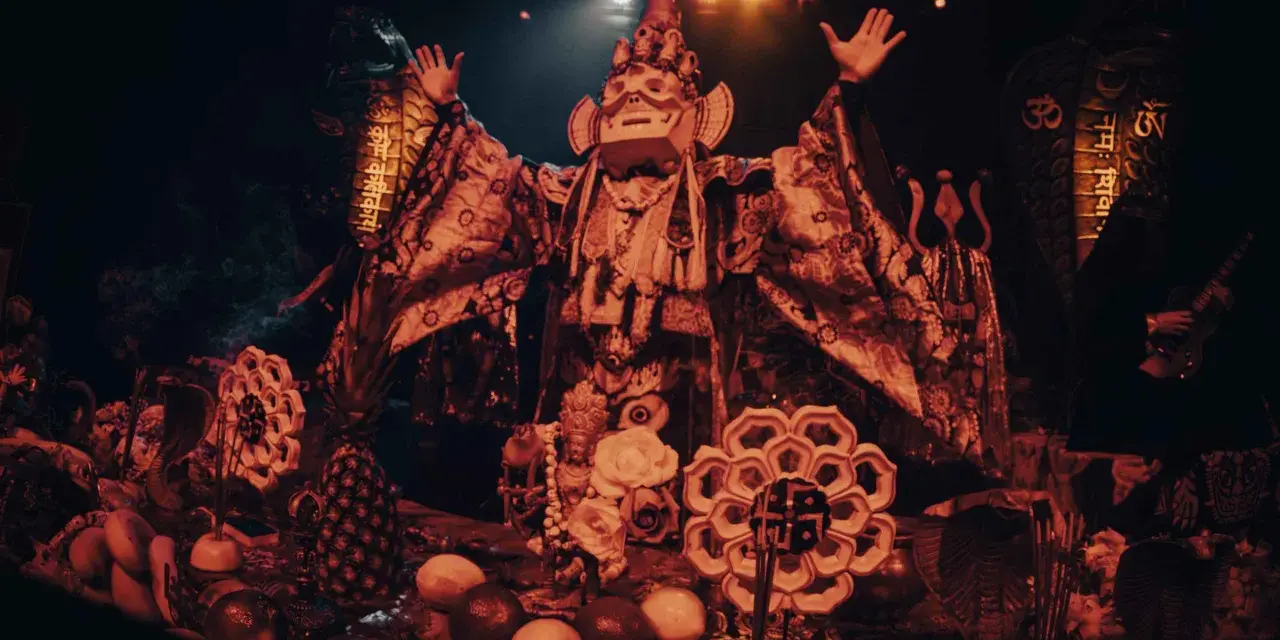 Un personnage masqué, drapé dans des tissus ornés, semble présider un rituel. Autour de lui, des offrandes de fruits, des serpents stylisés et des symboles sacrés évoquent un culte du feu mystérieux.
