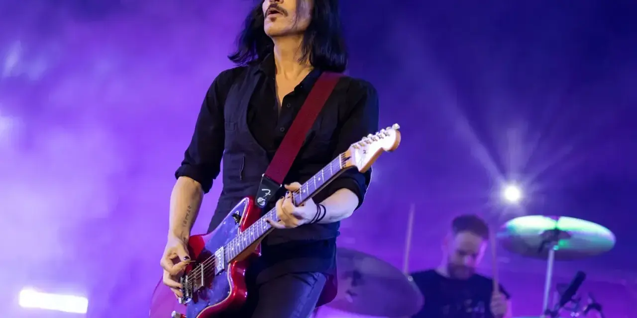 Brian Molko, le chanteur de Placebo, joue de la guitare rouge sur scène, éclairé par des lumières violettes.