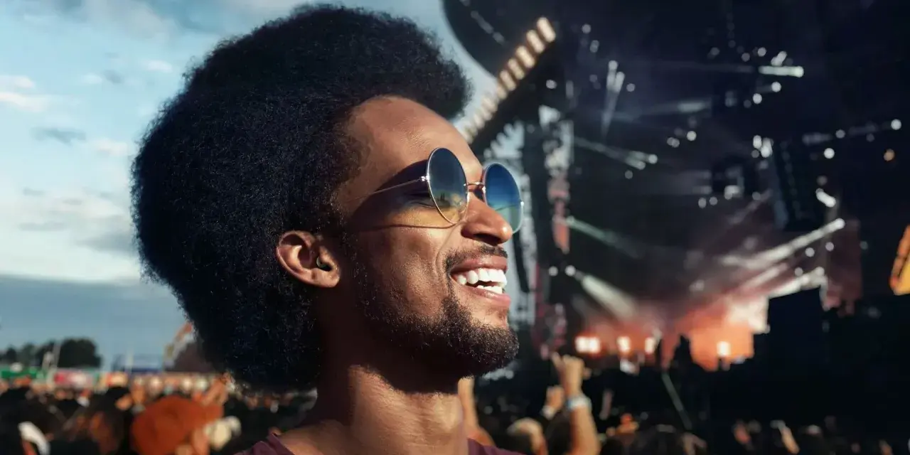 Un homme souriant avec des lunettes de soleil profite d'un concert en plein air. Il porte des bouchons d'oreilles, se demandant pourquoi pas de bouchon concert pour protéger son audition.
