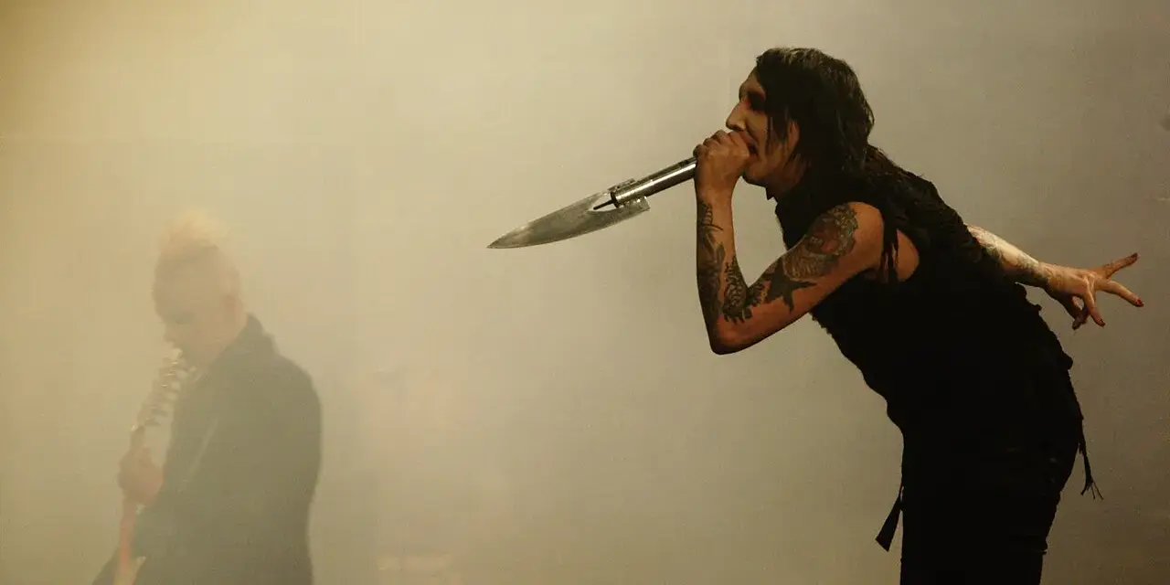 Marilyn Manson chante dans un micro en forme de couteau, un autre musicien est flou à gauche. L'ambiance d'un concert de Marilyn Manson album.