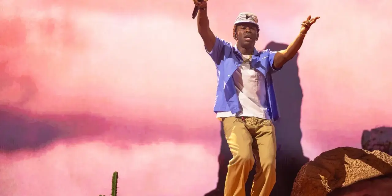 Tyler Okonma, micro en main, danse sur scène devant un décor rose pastel. Il porte une casquette, une chemise bleue ouverte sur un t-shirt blanc et un pantalon beige.