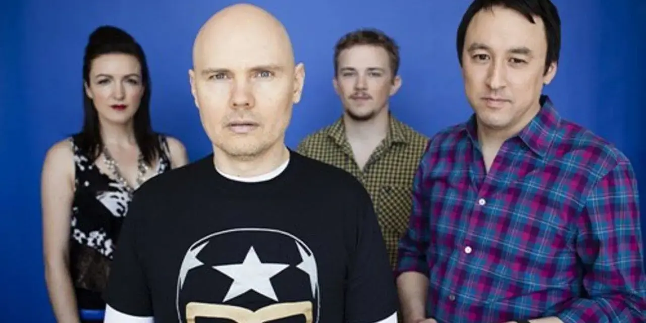 Les membres de The Smashing Pumpkins posent devant un fond bleu. Billy Corgan porte un t-shirt noir avec un masque de lucha.