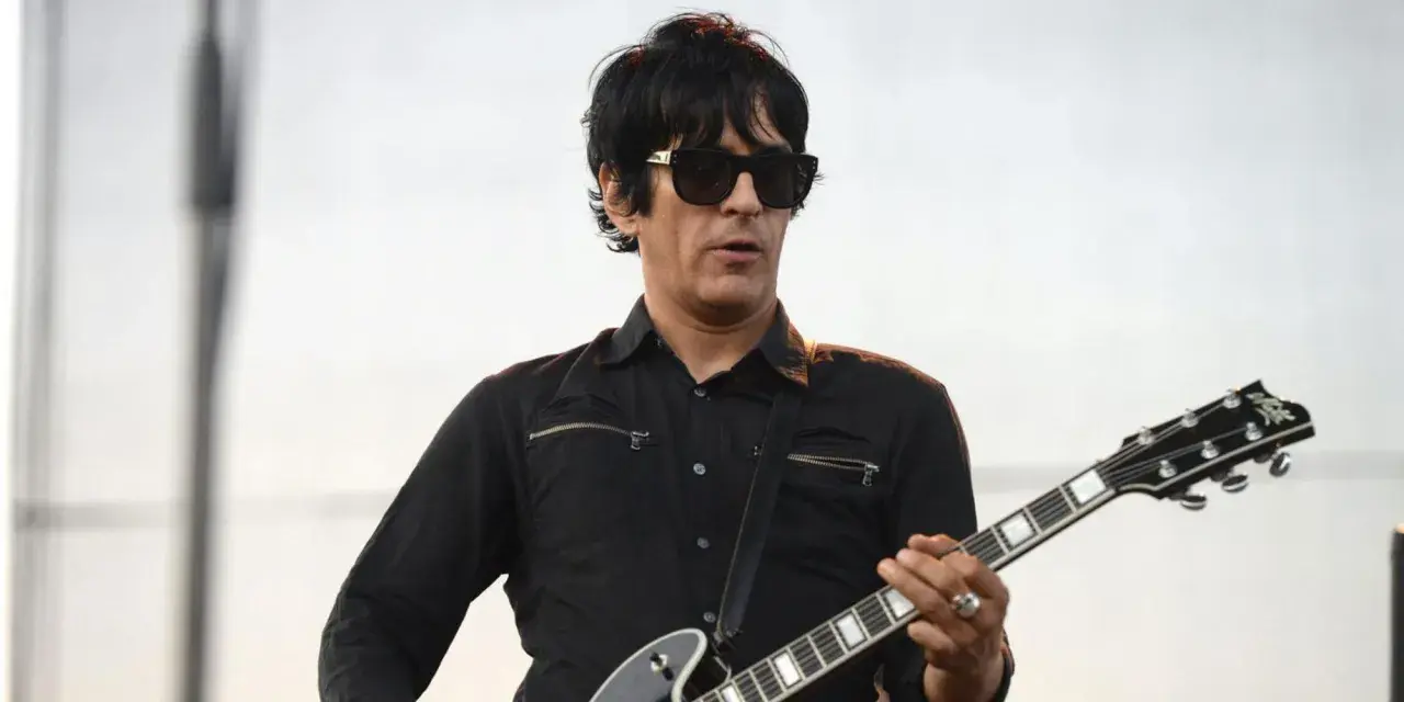 Un des membres de Eagles of Death Metal, guitariste en chemise noire et lunettes de soleil, joue sur scène.