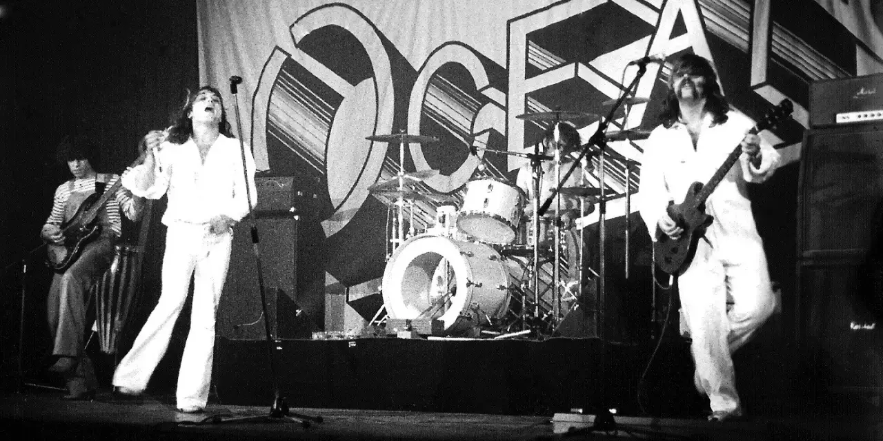 Le groupe Trust en concert, avec le chanteur en blanc au centre, un guitariste à gauche et un autre guitariste à droite. La scène est décorée d'une grande bannière avec le logo du groupe.