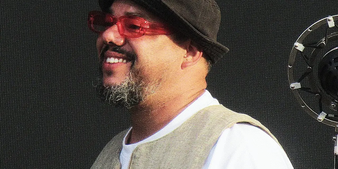 Un chanteur soul souriant, portant un chapeau et des lunettes rouges, se tient devant un microphone.