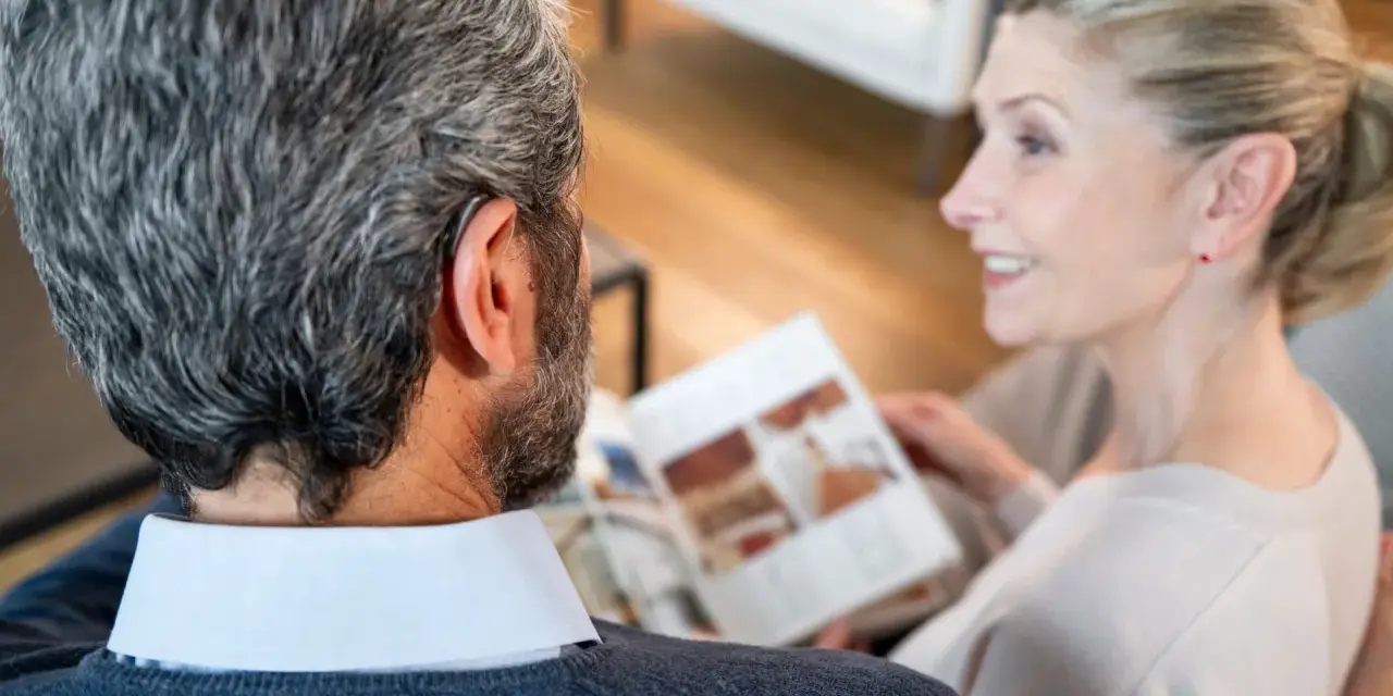 Un homme avec un appareil auditif, peut-être souffrant d'acouphènes après concert, discute avec une femme lisant un magazine.