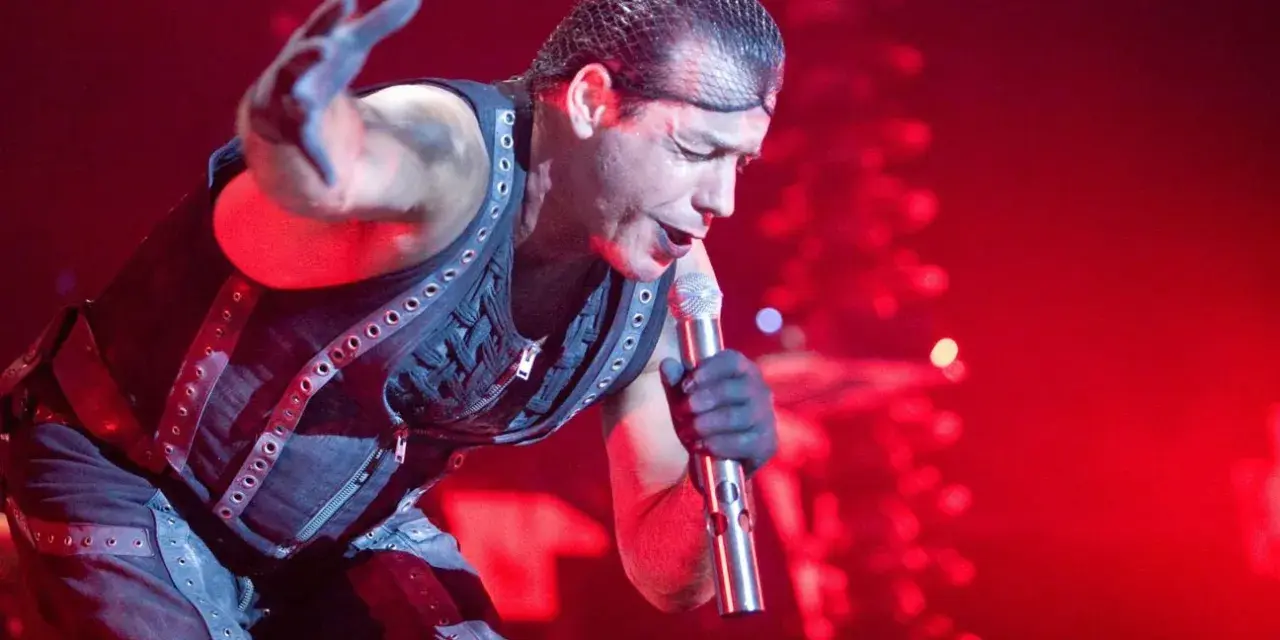 Till Lindemann, chanteur du groupe metal allemand Rammstein, en pleine performance sur scène, micro à la main, sous un éclairage rouge intense.