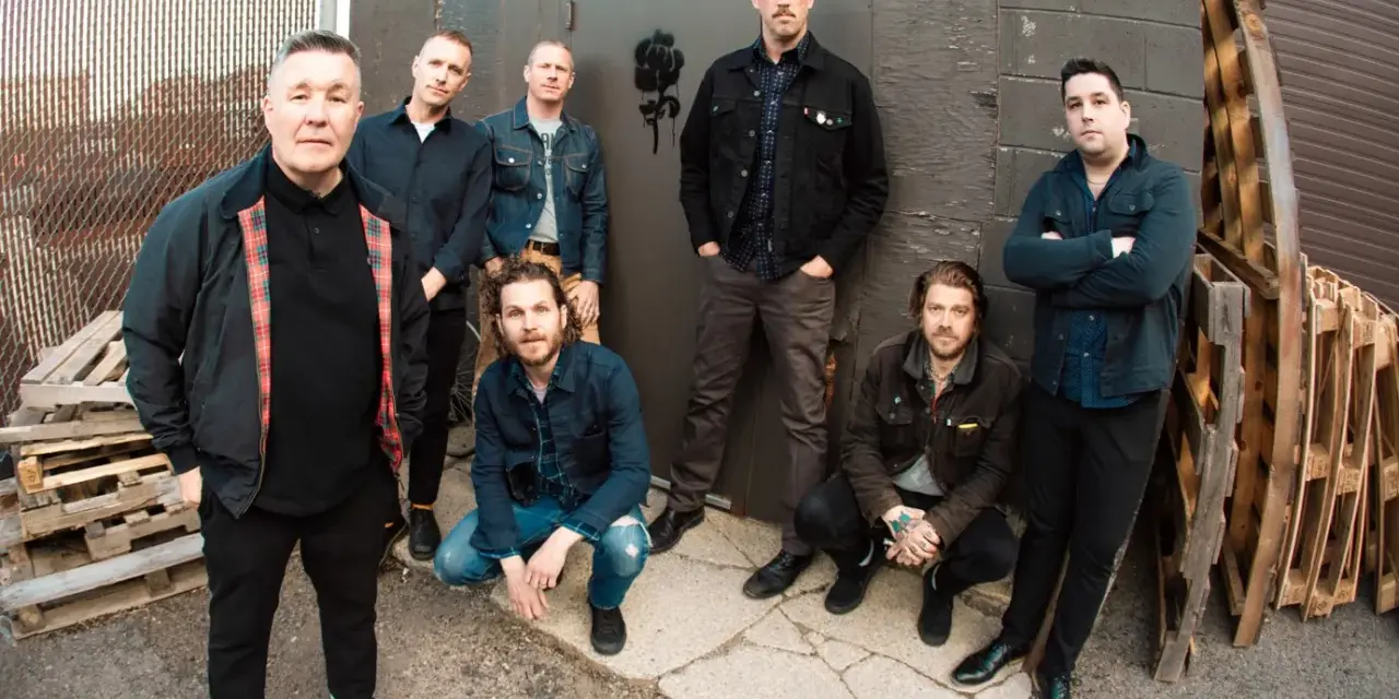 Les membres de Dropkick Murphys posent devant un mur sombre, certains accroupis, d'autres debout, prêts pour leur prochain concert.
