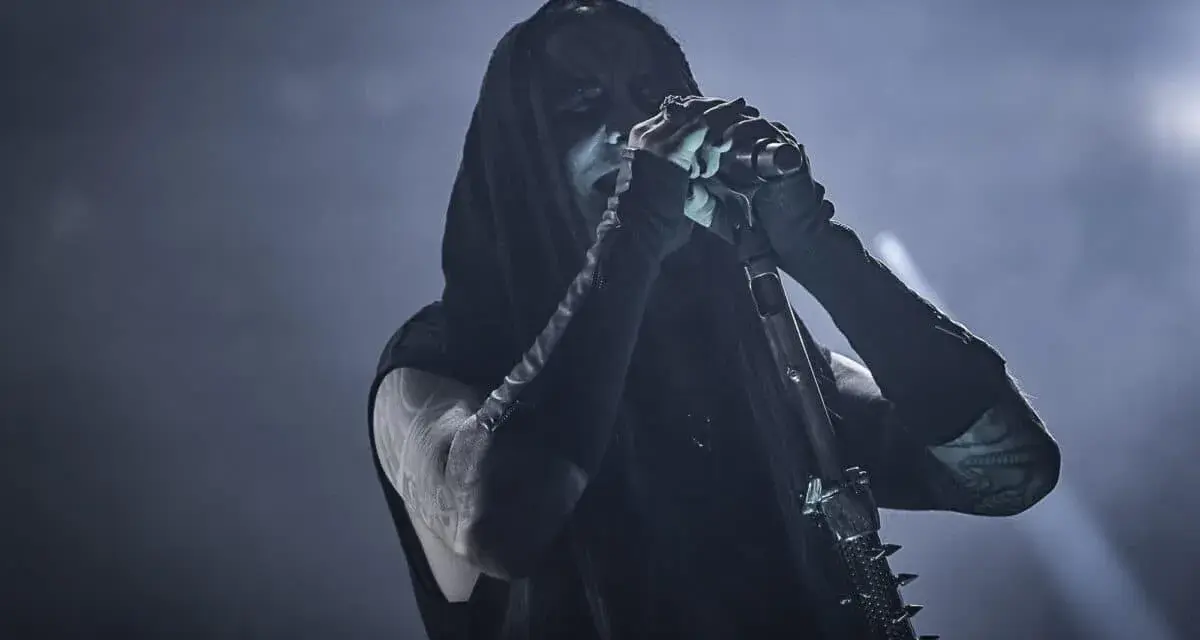 Chanteur de Dimmu Borgir, visage encapuchonné et maquillé, chantant dans un micro, ambiance sombre et enfumée.