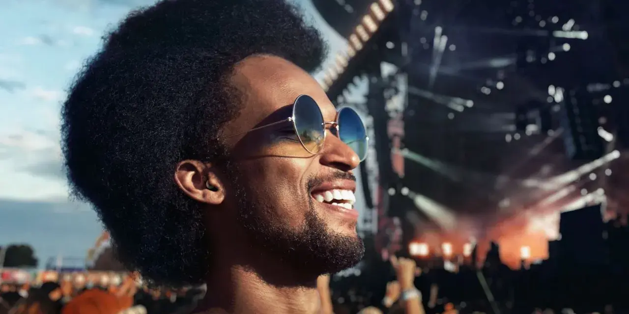 Homme souriant à un concert, portant des lunettes de soleil et un bouchon d'oreille pour protéger son audition.