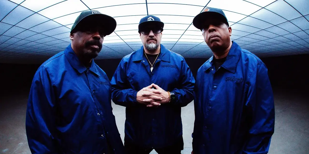 Les membres de Cypress Hill, B-Real, Sen Dog et DJ Muggs, posent ensemble, portant des vestes bleues assorties sous un plafond géométrique.