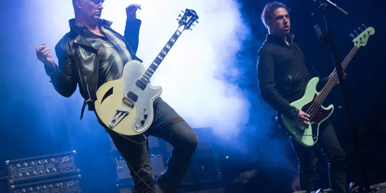 Deux membres de Queens of the Stone Age en pleine performance, l'un jouant de la guitare, l'autre de la basse, sous des éclairages bleus intenses.